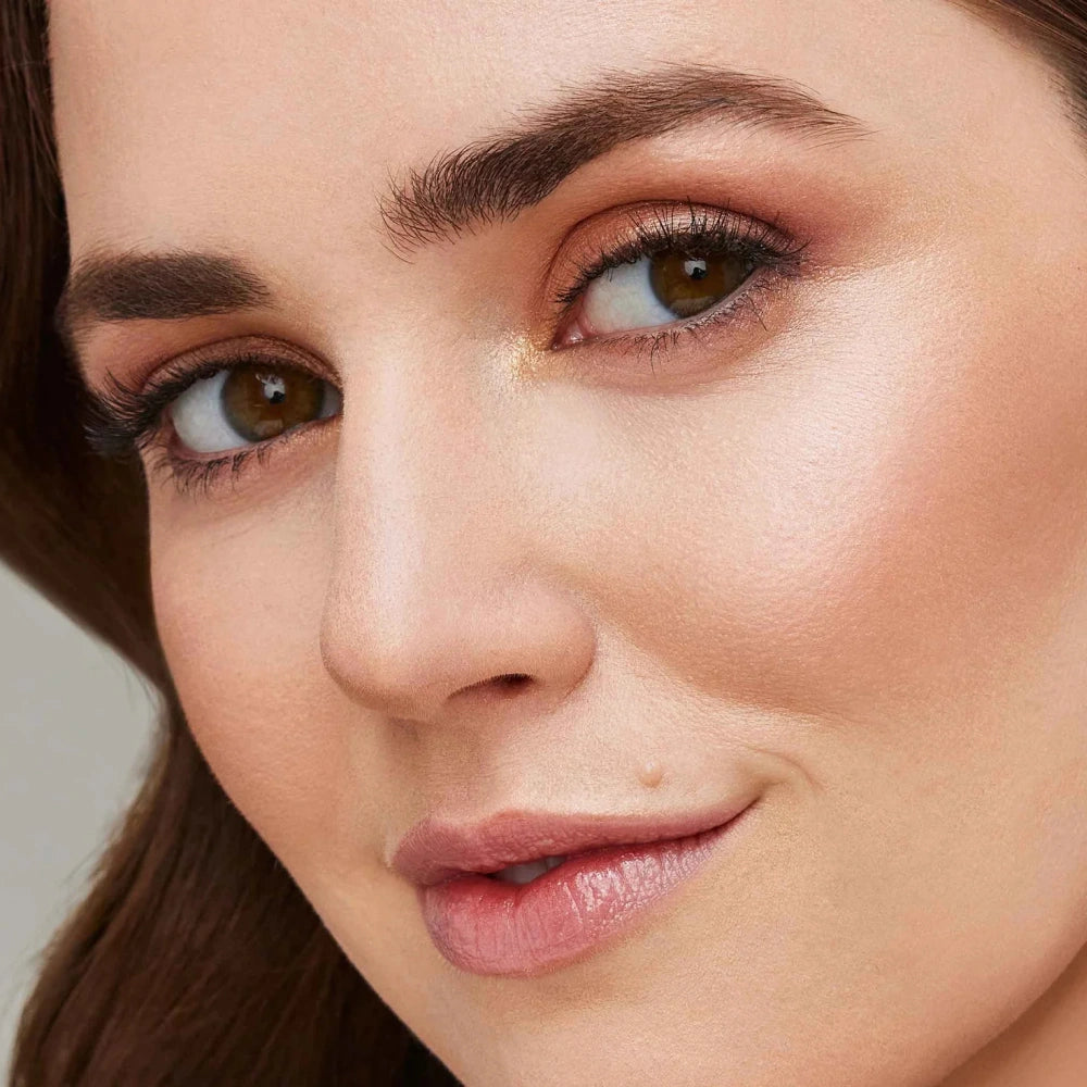 Close-up of a woman's face showing a shimmering gold eyeshadow on her eyelid, with warm pink lipstick and defined brown eyebrows.