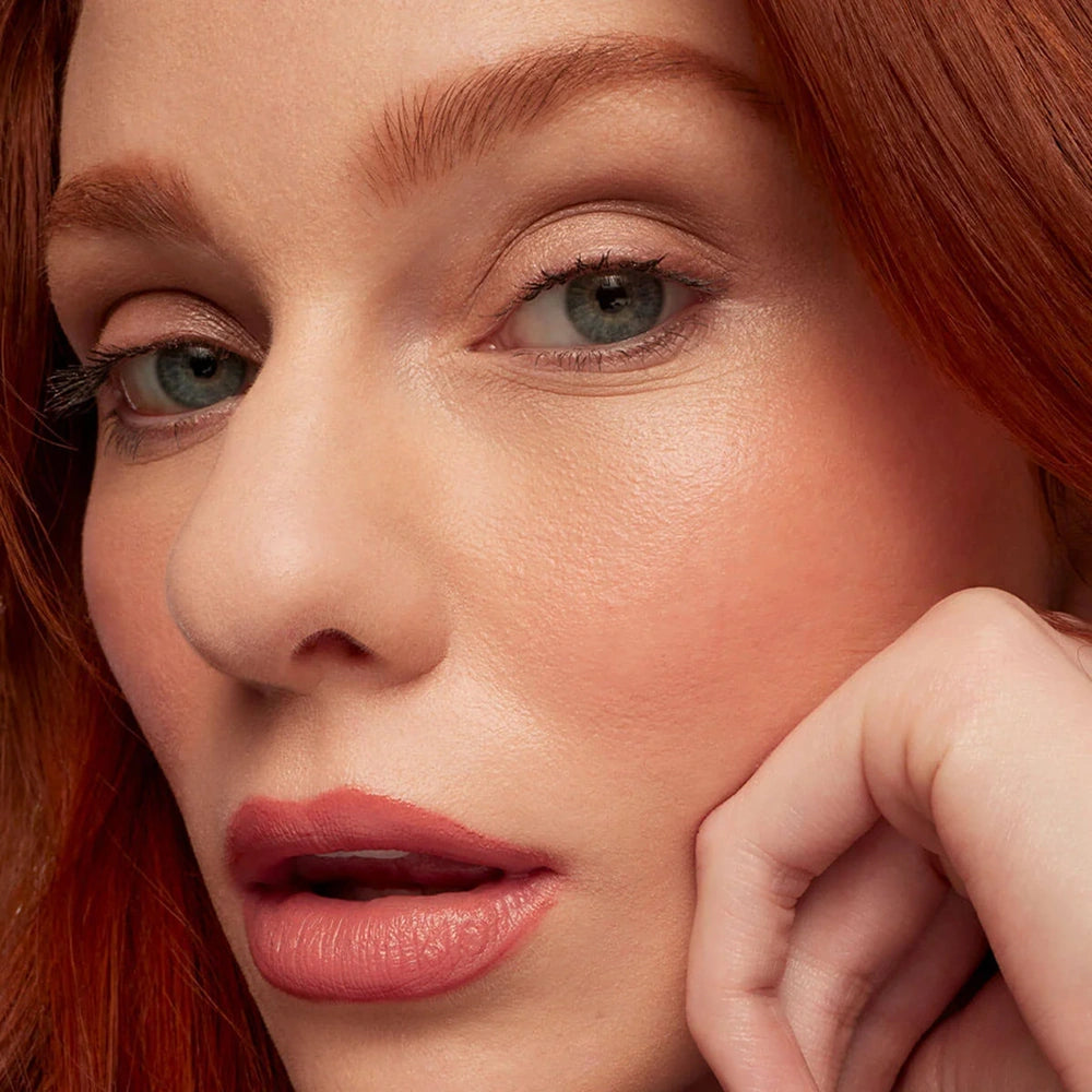 Close-up of a woman with red hair and green eyes wearing a glossy pink lipstick, resting her chin on her hand.